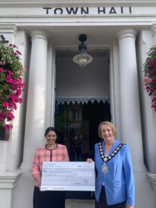 Standing on the steps of Witham Town Hall, Priti Patel MP hands a cheque to Town Mayor, Cllr Angela Kilmartin for her chosen charities, MacMillan Cancer Support and the Witham Amateur Operatic Society.